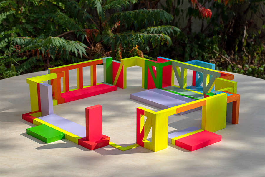 An architectural mock-up. Colorful, geometric design elements are arranged in a square within a courtyard. Trees are in the background.