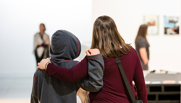 Two people viewed from behind stand in the galleries with their arms around each other. Two artworks hang on a white wall in the blurred background.