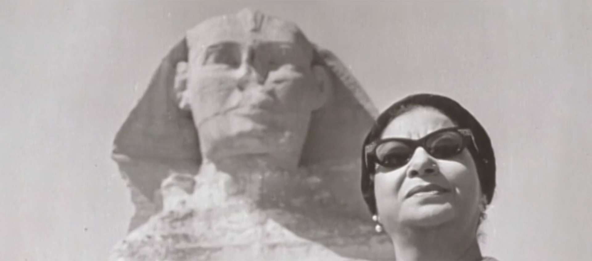 A black-and-white photo of Umm Kulthum, who wears dark sunglasses and a dark hat. Behind her is the Great Sphinx of Giza.