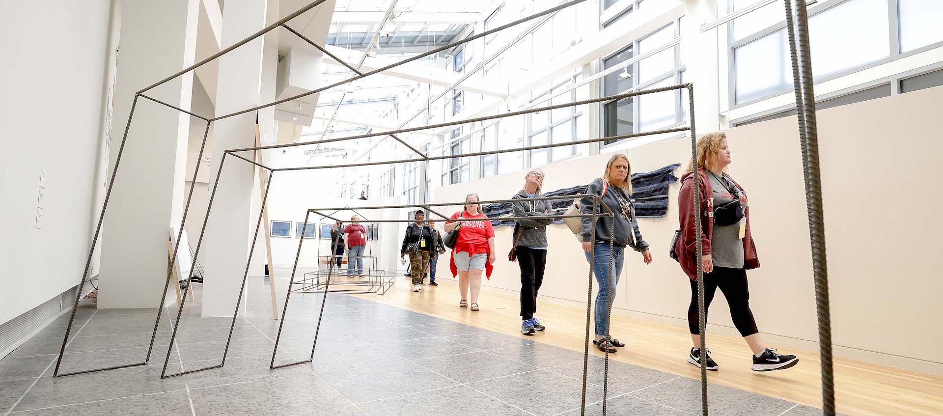 A line of seven people walk through a gallery space. A sculpture made of tiered rebars is in the foreground.