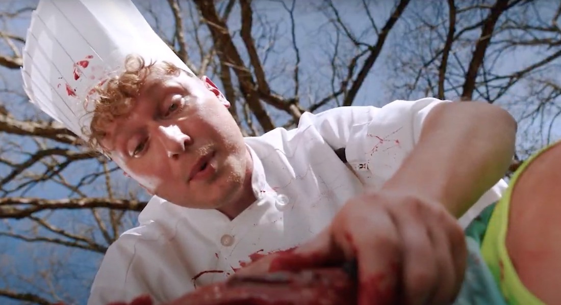 A young man in a chef outfit looks down and holds a bloody head, which is mostly out of frame.