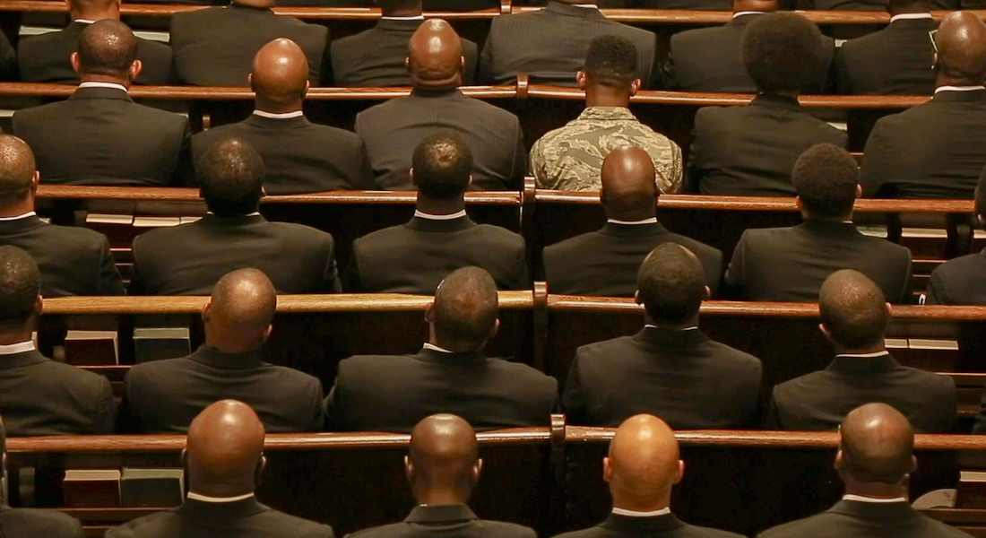 Several people in dark suits, one in military fatigues, sit in pews.