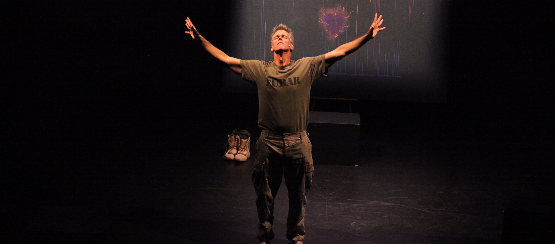 A man in army fatigues stands on a stage with his head back and his arms extended outward.