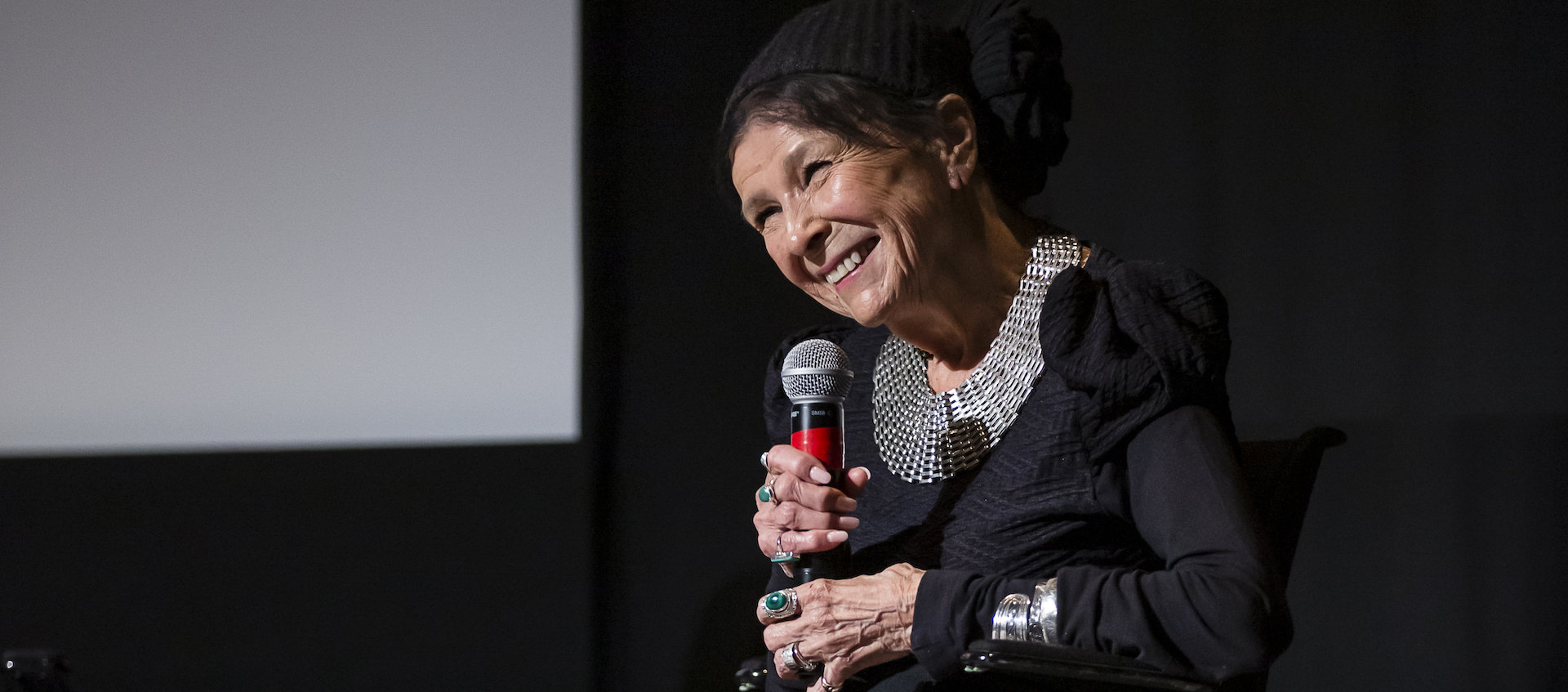 An older woman wearing black clothing and abundant silver jewelry sits on a stage and smiles as she holds a microphone.