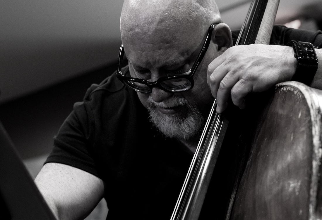 A bald man with a gray beard and glasses looks down as he plays a standup bass.