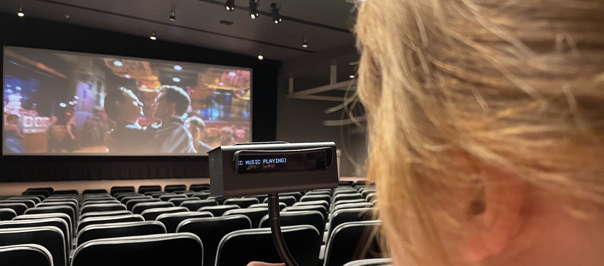 Woman sits in a movie theater and uses a hand-held device to see close captioning for a film on screen.