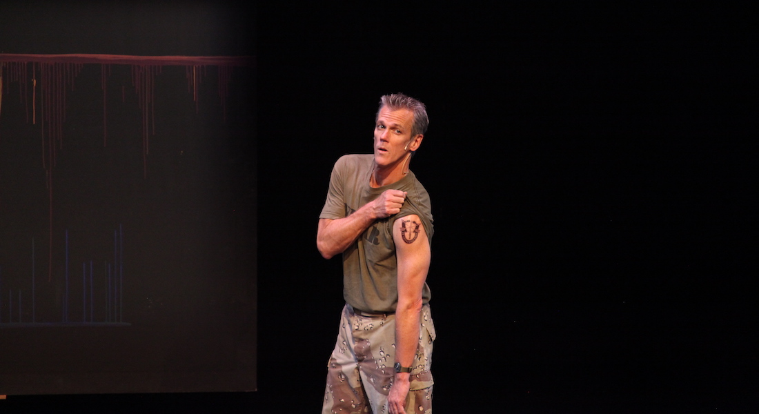 A man stands on a darkened stage in military fatigues and holds up one sleeve to show a tattoo
