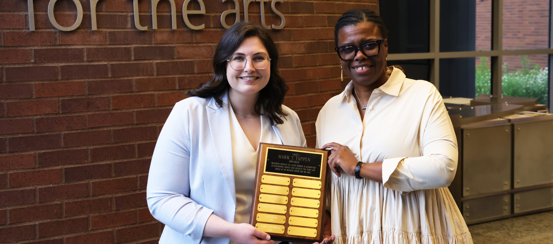 Two women stand against a brick wall smiling and holding an award plaque.