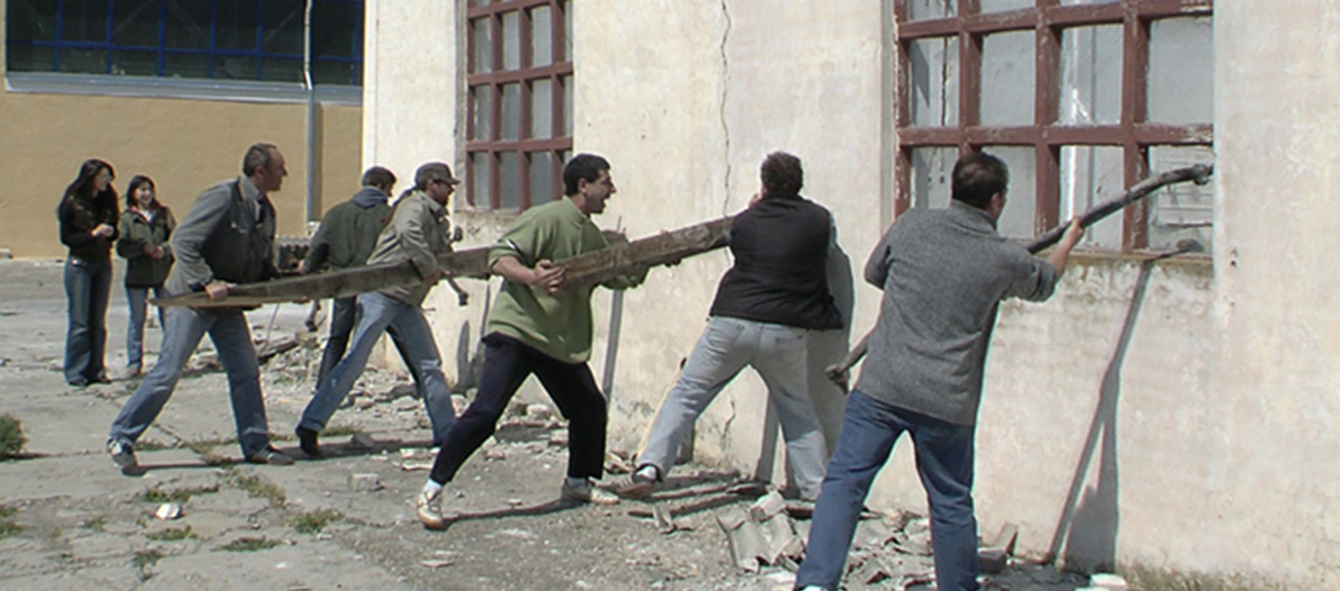 A row of men use logs and other objects to knock down the wall of a decaying building.