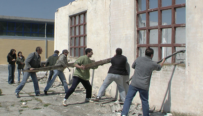 A row of men use logs and other objects to knock down the wall of a decaying building.