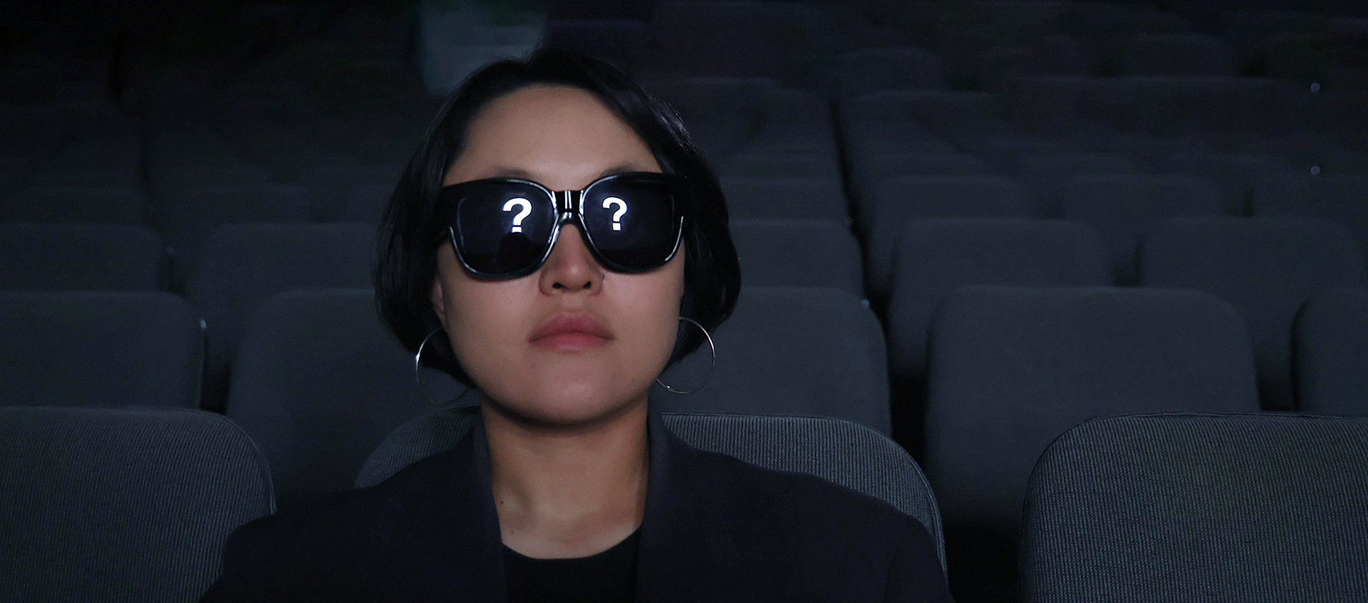 A woman with short black hair sits in a dark theater with dark glasses on, there are question marks in reflecting in the lenses of the glasses.
