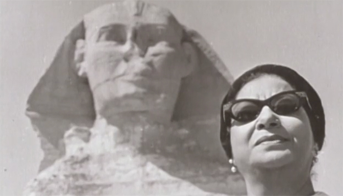 A black-and-white photo of Umm Kulthum, who wears dark sunglasses and a dark hat. Behind her is the Great Sphinx of Giza.