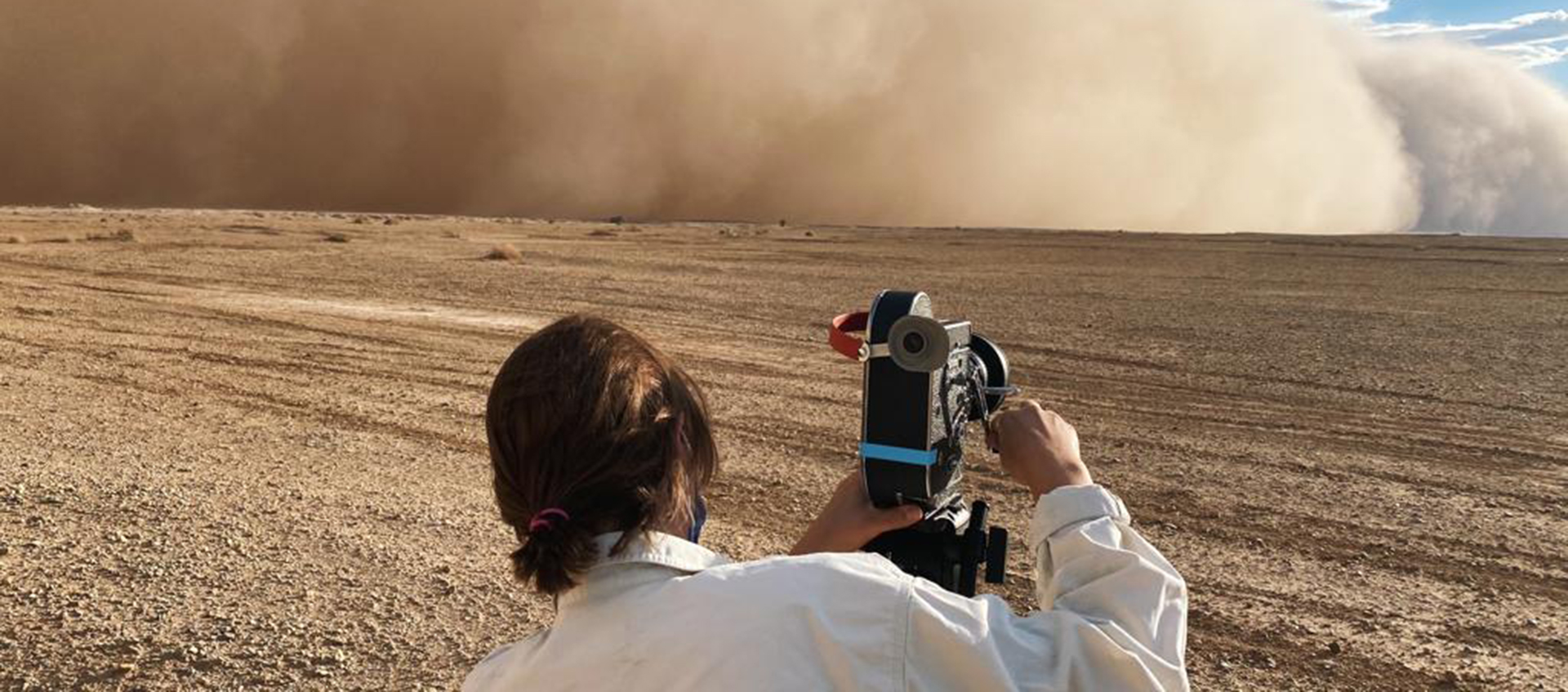 Director Terra Long holds a 16mm camera. She wears a white shirt and faces a dust storm.