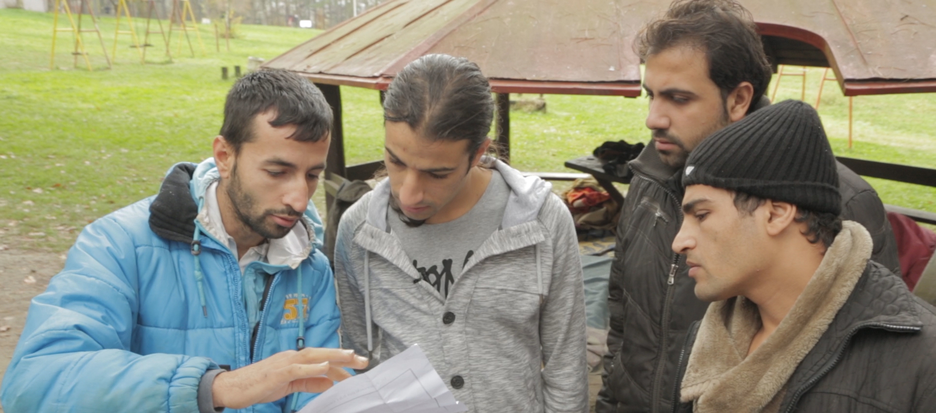 Four Serbian men stand close together to all look at a piece of paper in a field next to a pavilion