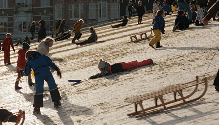 A large group of children in winter clothes sled down a snowy hill with urban buildings visible behind them. 