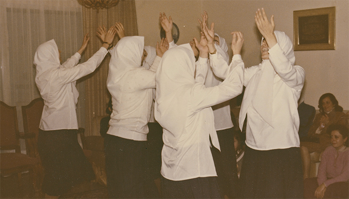 An archival photo of a group of six women all wearing white hijabs and blouses with their arms raised towards the ceiling. 