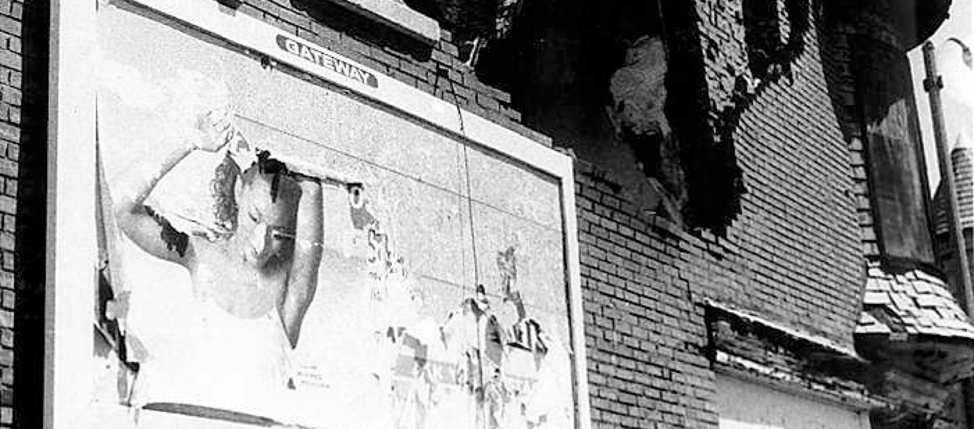 A black and white image of the front of a dilapidated brick building and a deteriorating billboard. 