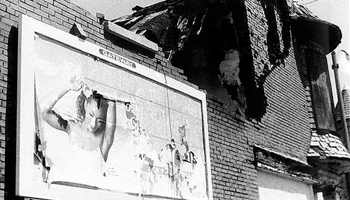 A black and white image of the front of a dilapidated brick building and a deteriorating billboard. 
