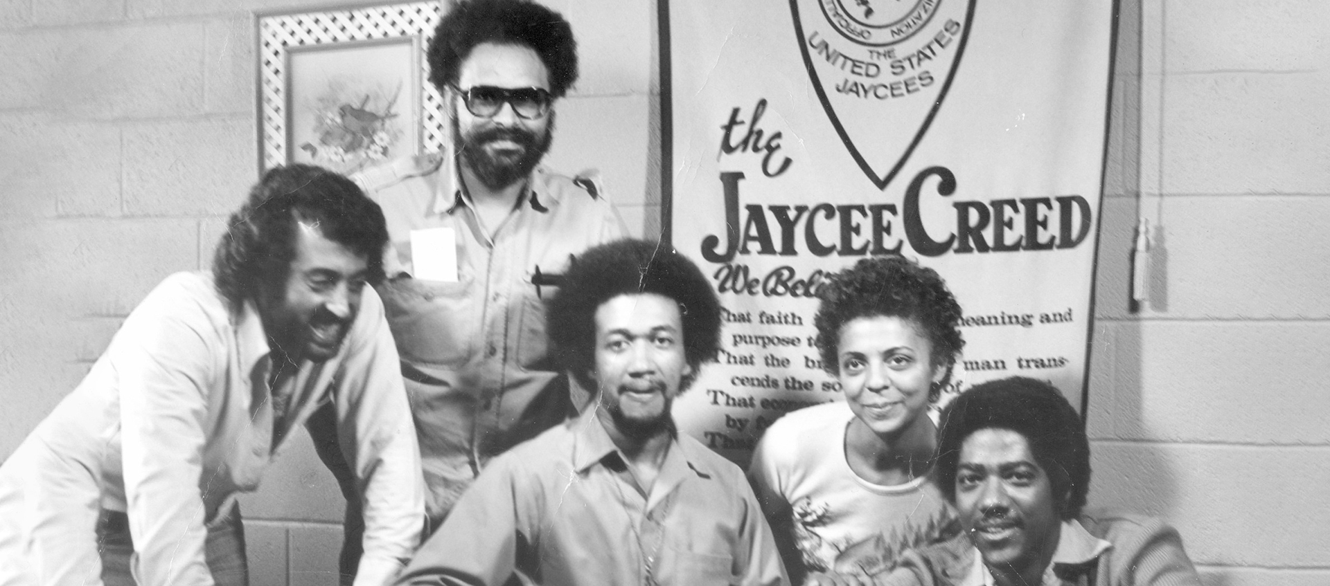 A black and white photo of five Black people sitting and standing in front of a banner that reads “the Jaycee Creed.".” 