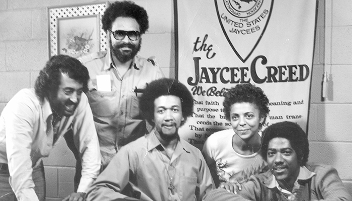 A black and white photo of five Black people sitting and standing in front of a banner that reads “the Jaycee Creed."