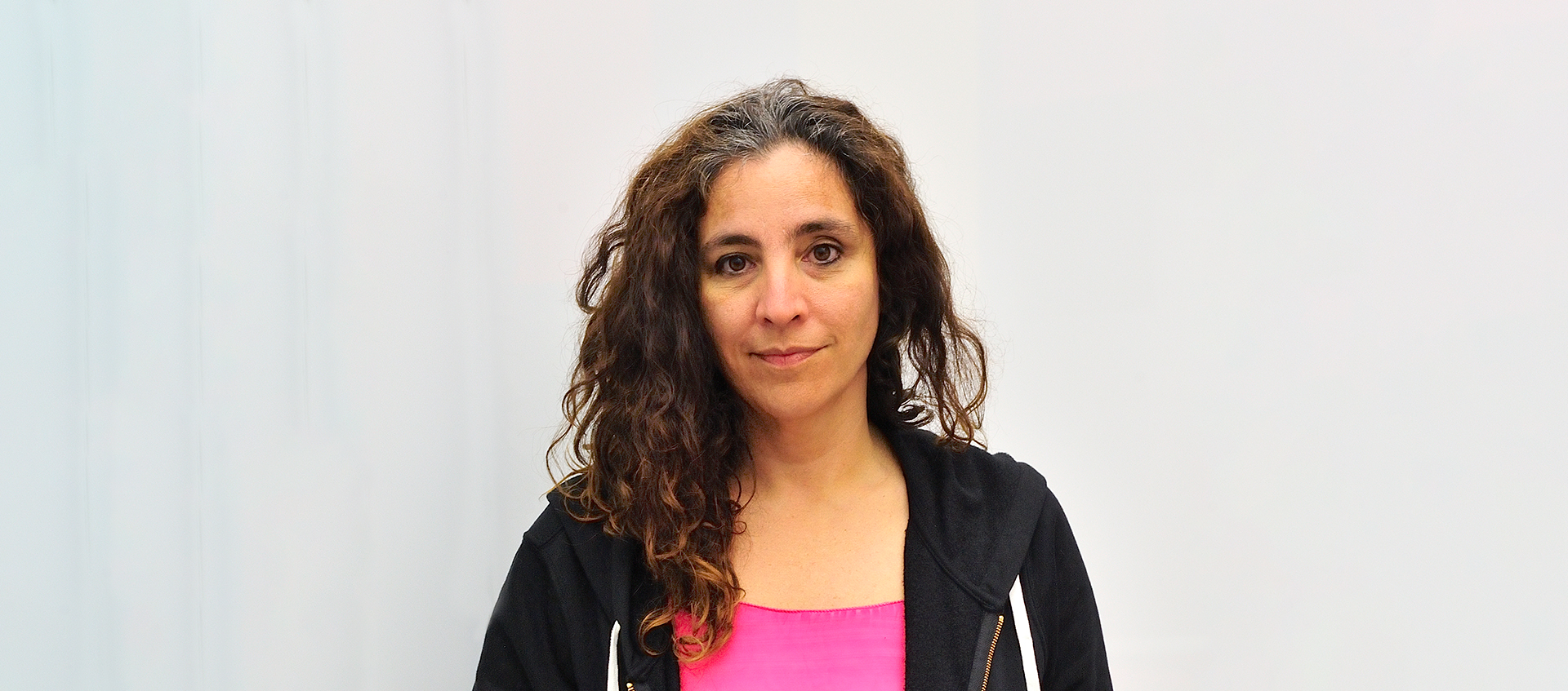 A photograph of Beatriz Santiago Muñoz who has long brown hair, wears a black zip hoodie with a pink shirt, against a white background.