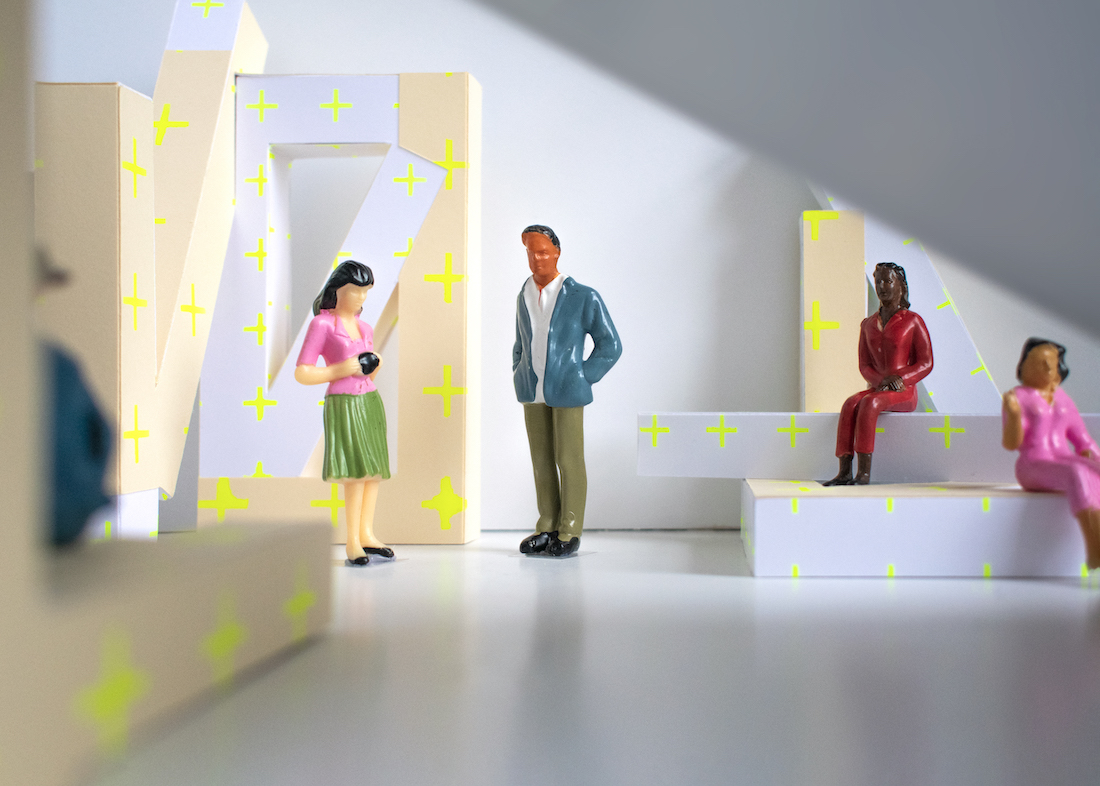 Detail of figurines in architectural space. The figurines interact with the architectural elements. The architectural elements are geometric with neon yellow plus signs on them. 