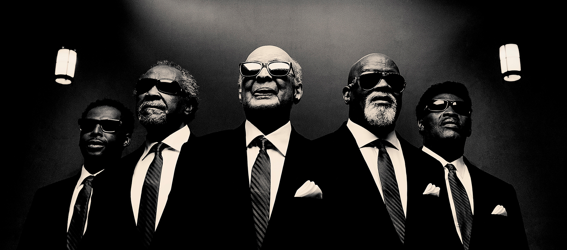 Black-and-white photo, shot from a low angle, of five older Black men wearing suits, ties, and sunglasses. There is a rose window above them.