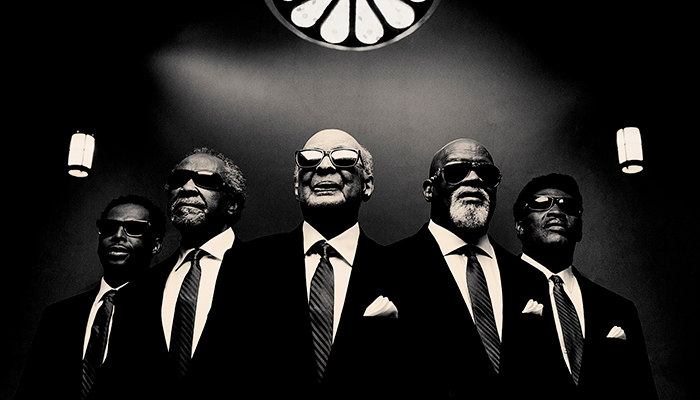 Black-and-white photo, shot from a low angle, of five older Black men wearing suits, ties, and sunglasses. There is a rose window above them.