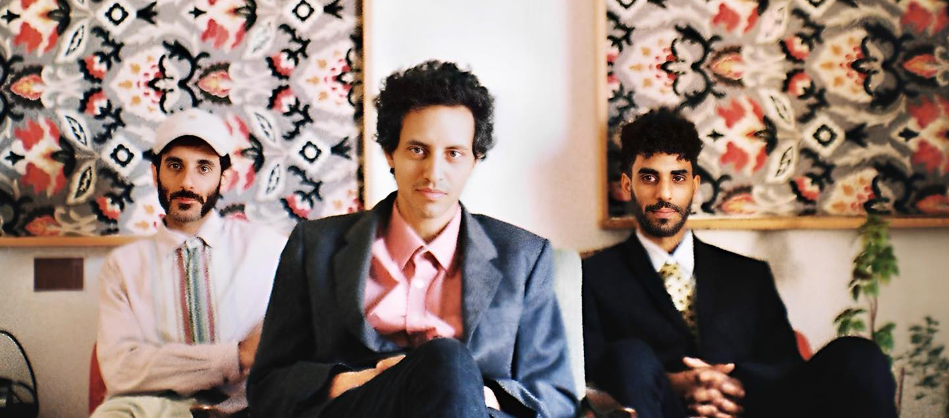 Three members sit in chairs in front of two large frames featuring red, black, and white patterns. Band leader Eyal El Wahab sits in the forefront.