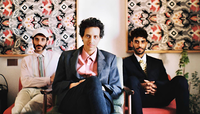 Three members sit in chairs in front of two large frames featuring red, black, and white patterns. Band leader Eyal El Wahab sits in the forefront.
