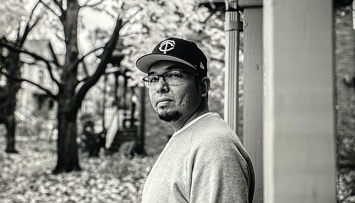 Black-and-white photo of Joe Rainey, who has a dark goatee and wears glasses and a baseball cap. He stands outside under a porch roof.