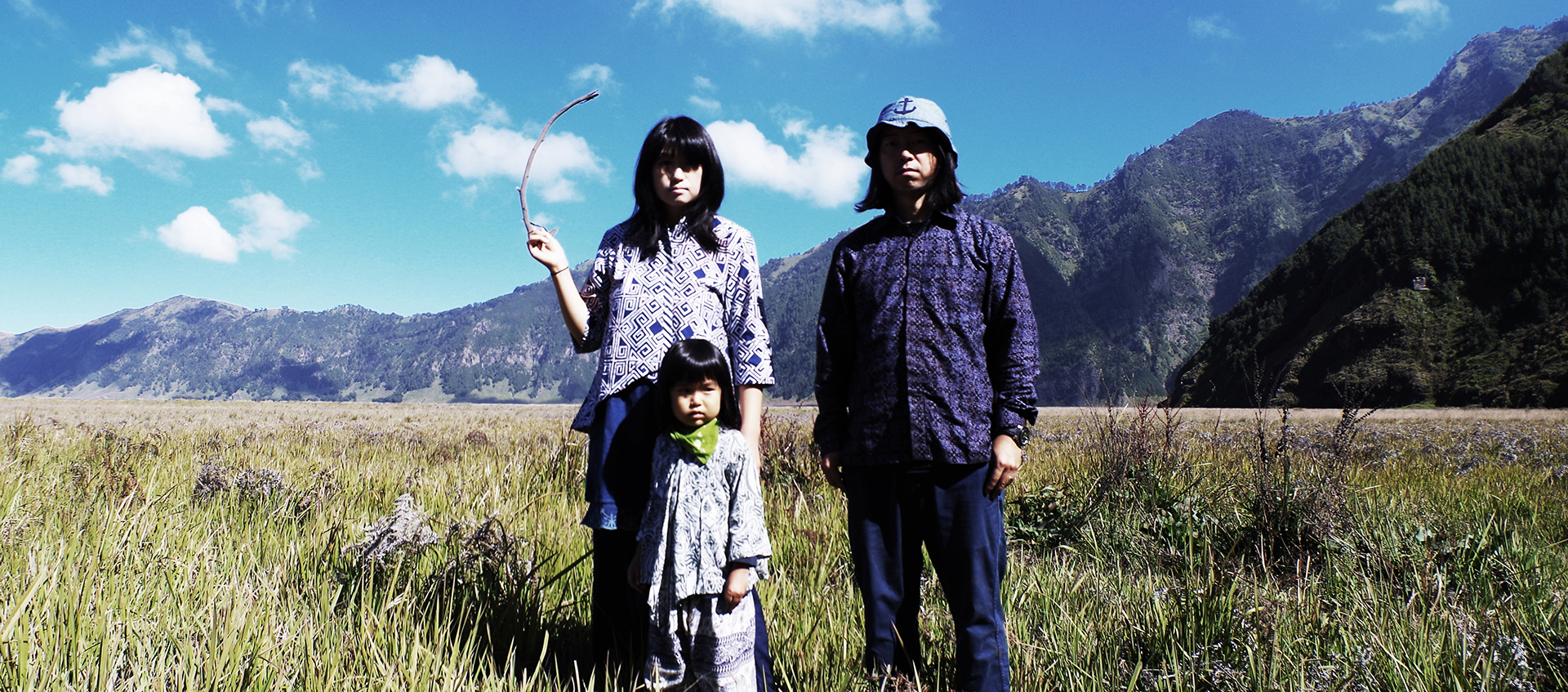 Band members itta and Marqido and their young child stand in a grassy field. There are mountains and a blue sky filled with clouds in the background.