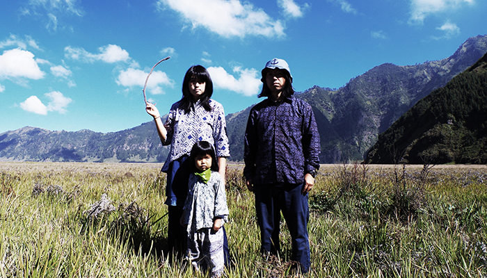 Band members itta and Marqido and their young child stand in a grassy field. There are mountains and a blue sky filled with clouds in the background.