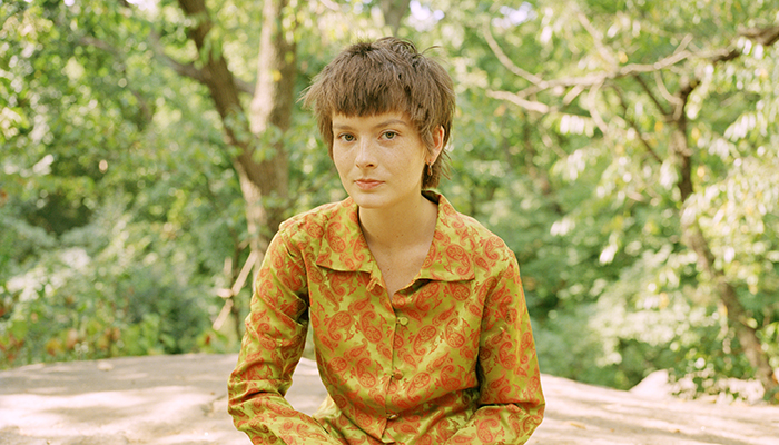 Zoh Amba sits in a forest. She wears a yellow and orange paisley-patterned shirt and has short, light brown hair and white skin with freckles.