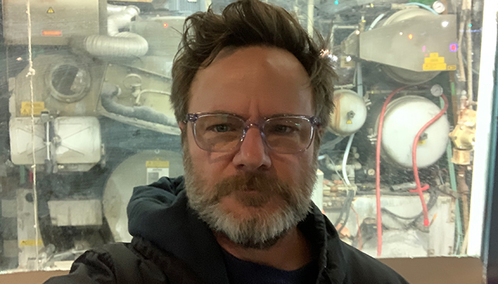 A man with a gray beard and clear-framed glasses stands in front of heavy industrial equipment.