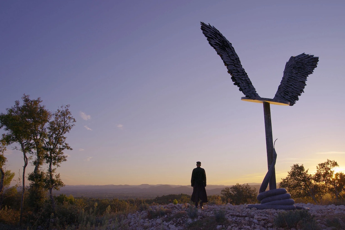 A man stands next to a large, tall sculpture of wings against a wide expanse at sunset.