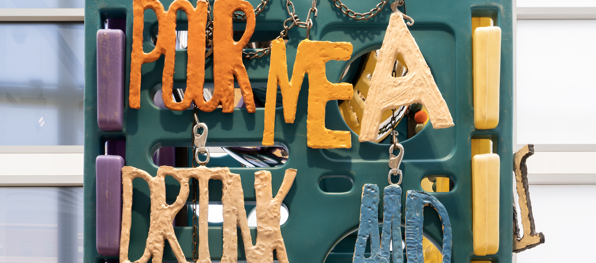 A detail of a colorful sculptural work with hand wrought lettering that spells out “Pour me a drink and...”