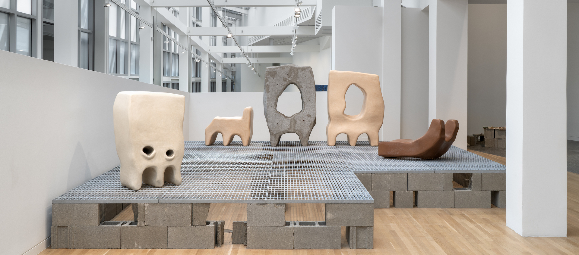 A gallery space holds five sculptures of various size, all sitting on metal grating and cinder blocks.