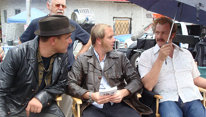Three men sit in director's chairs looking at each other, one is holding an umbrella and another is wearing a fedora.