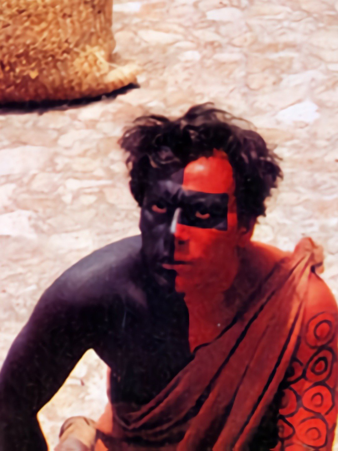 A person with short dark hair and red and black full body paint looks up toward the camera.