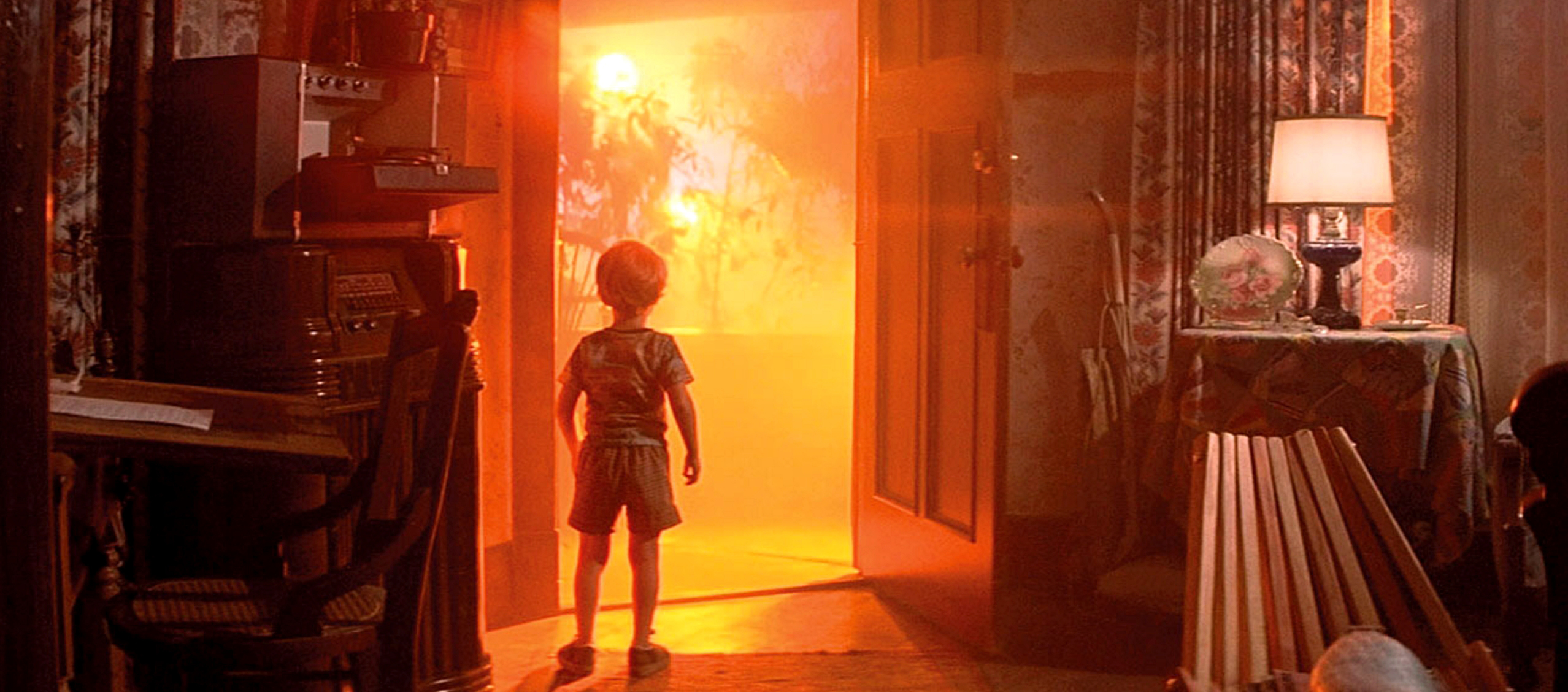 A little boy is standing in an open doorway, from which an orange light emanates.