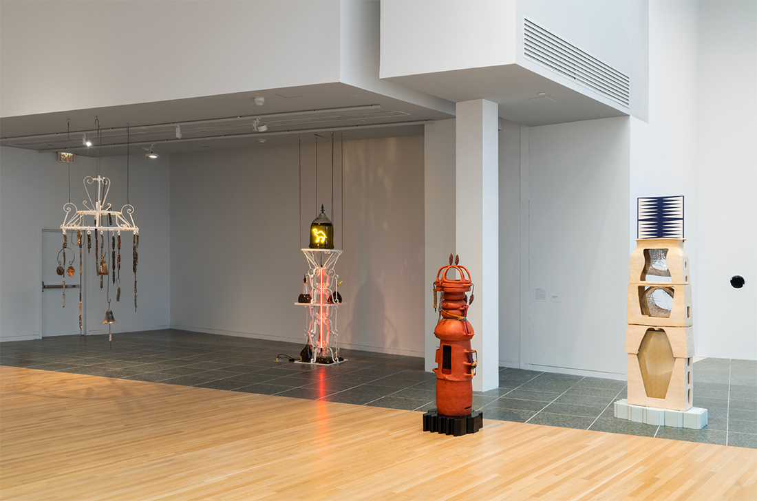 Installation of four sculptures made of metal and ceramics resting on a divided wood and granite floor. White beams and soffit are visible.