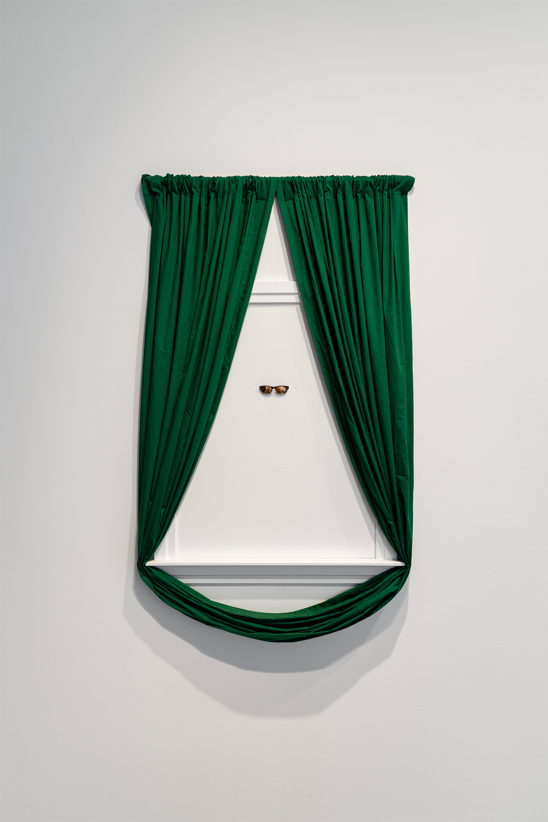 Green curtains surround a white window frame attached to the wall. A pair of sunglasses is affixed to the wall, roughly centered in the frame.