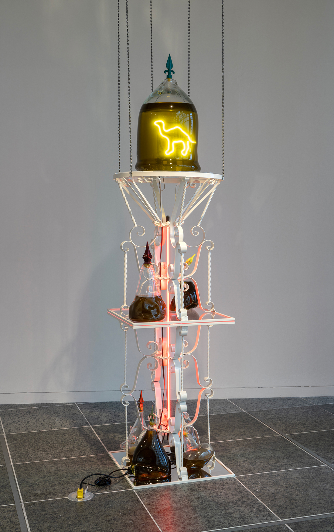 Three-level sculpture of conjoined metal tables. Camel neon light in glass vase full of olive oil at top. Olive oil bottles rest on the tables.