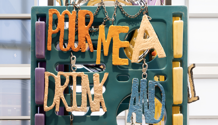 A detail of a colorful sculptural work with hand wrought lettering that spells out “Pour me a drink and...”