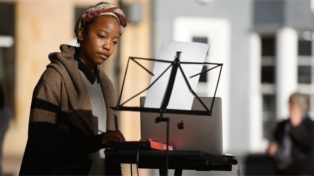 A woman wearing a sweater looks at papers on a music stand while turning the knobs on a panel next to a laptop. 