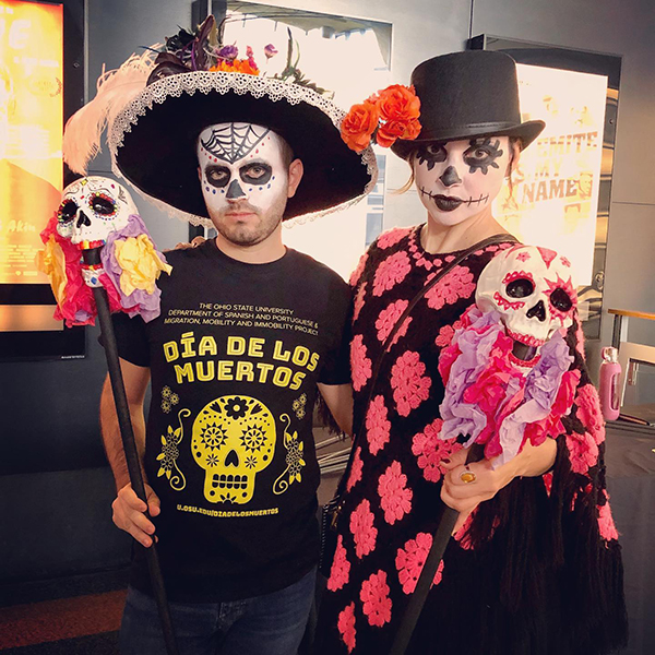 Two people wearing black-and-white skull face paint, a sombrero and top hat, and festive shirts, are carrying canes topped with decorative skulls.
