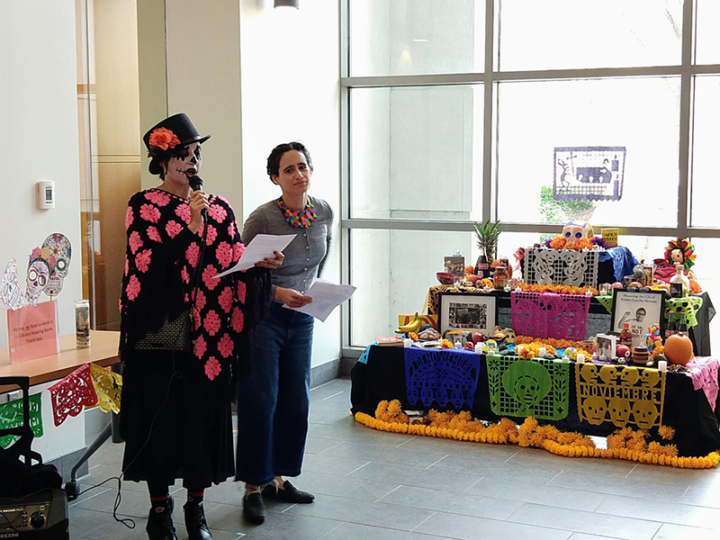 Two people stand next to an ofrenda. One is wearing a black and pink shawl and top hat and holding a microphone while speaking. 