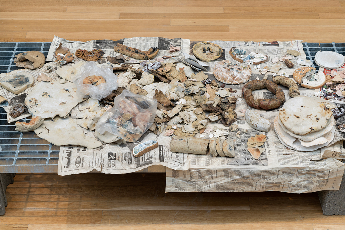 Overhead of ceramic sculptures made to resemble small, multicolored breads in various states of decomposition. They are placed on newspapers.