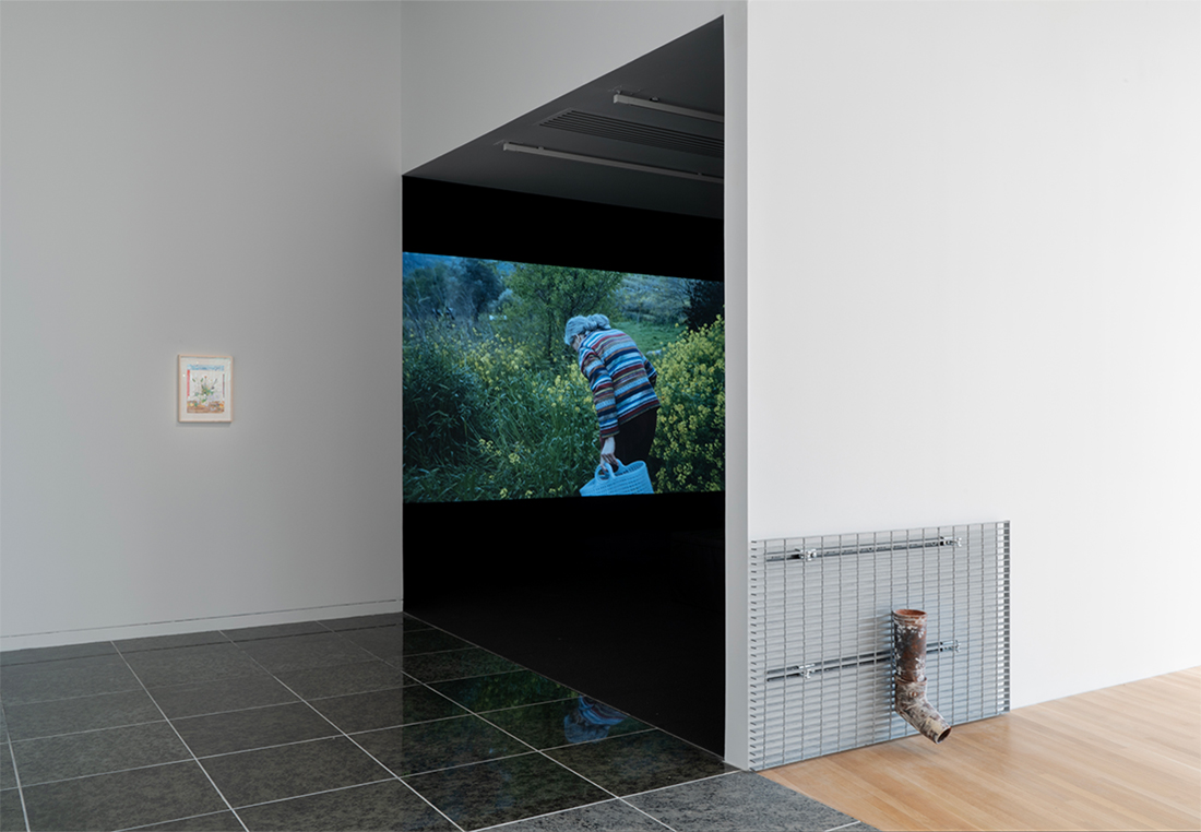 View of entrance to a dark room with video playing. A wall work is on left-middle of wall and a sculpture on a metal grate is on the bottom right. 
